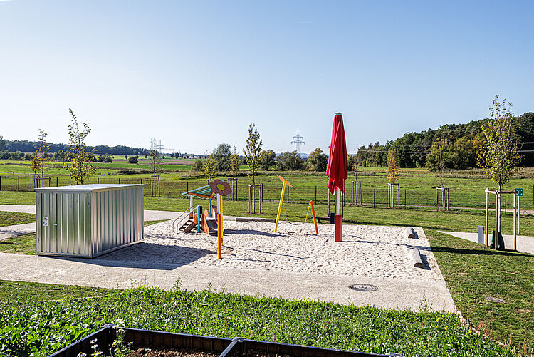 Sandspielbereich der Kita mit Spielgerät, Sonnenschirm und weitläufiger Grünfläche