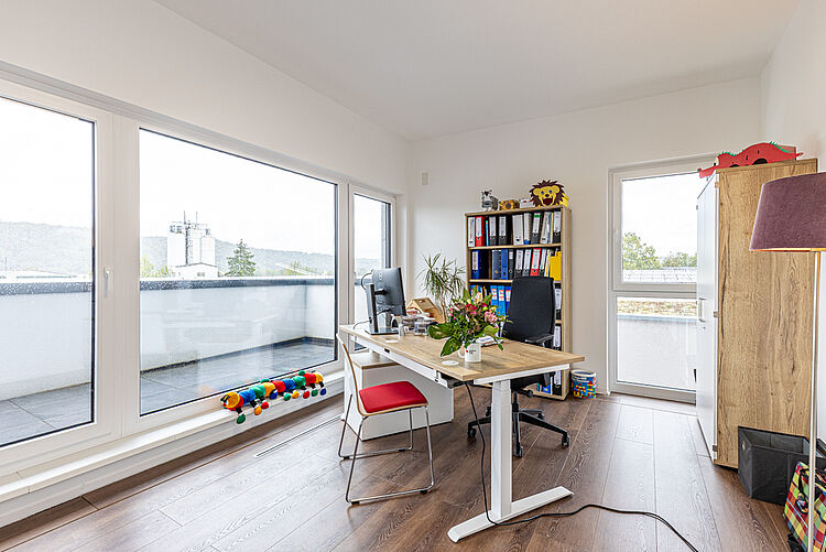 Schreibtisch mit Blumenstrauß und PC in einem hellen Raum mit großer Fensterfront