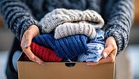 Hände halten einen Karton mit sortierten, bunten Strickpullovern für den DRK Kleiderladen