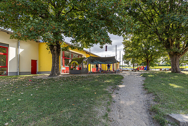 Kita-Gebäude mit Spielplatz, Sonnensegel und schattenspendenden Bäumen