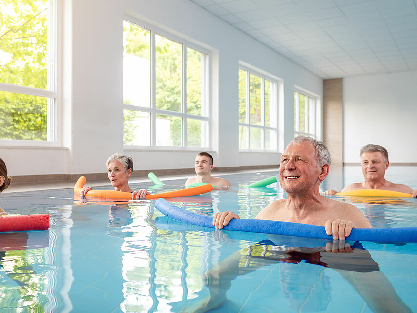 Gruppe von Menschen macht gemeinsam Wassergymnastik mit Poolnudeln in einem hellen Schwimmbad