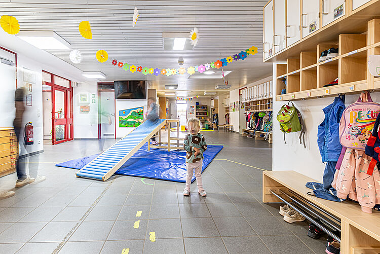 Mädchen spielt im Eingangsbereich der Kita „Rabennest“ in Rabenau-Londorf, umgeben von bunten Jacken und Rucksäcken