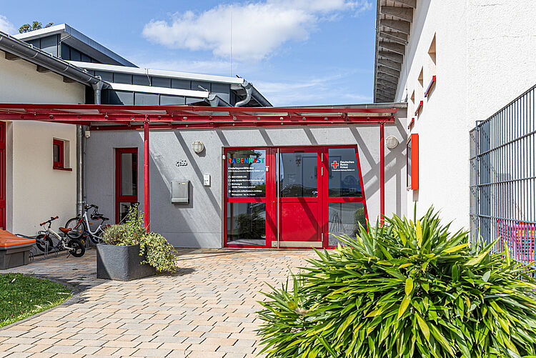 Eingangsbereich der Kita mit roter Tür, Beschilderung und Fahrradständern