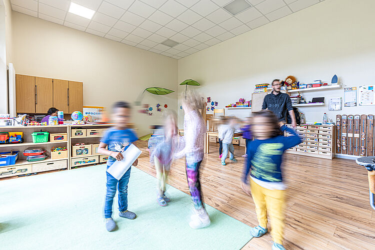 Kinder spielen und bewegen sich im Gruppenraum mit Lernmaterialien und Erzieher