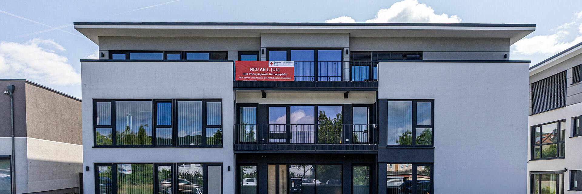 Modernes Gebäude der neuen DRK Logopädiepraxis in Marburg-Cappel mit Banner „Neu ab 1. Juli“