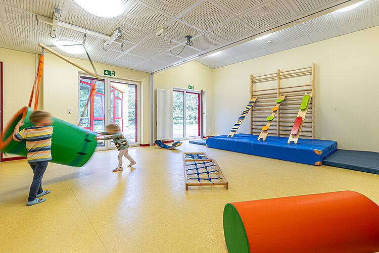 Kinder spielen im Bewegungsraum der Kita mit Klettergeräten, Matten und bunten Elementen