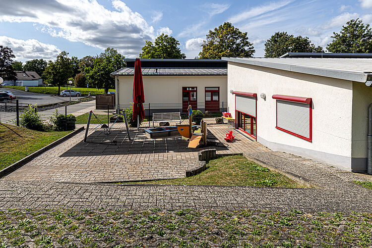 Blick auf die Spielterrasse mit Sandkasten, Sonnenschirm und Rutschfahrzeugen