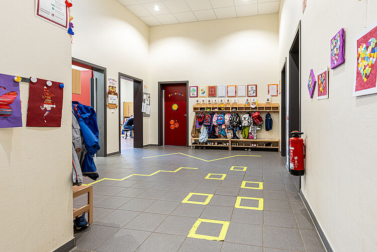 Blick in den Flur der Kita mit Kinderkunstwerken, Garderoben und bunten Markierungen am Boden