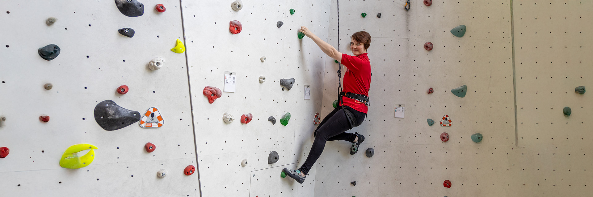 Eine Frau in rotem DRK-T-Shirt klettert lächelnd an einer hellen Kletterwand mit bunten Griffen – therapeutisches Klettern beim DRK Mittelhessen.